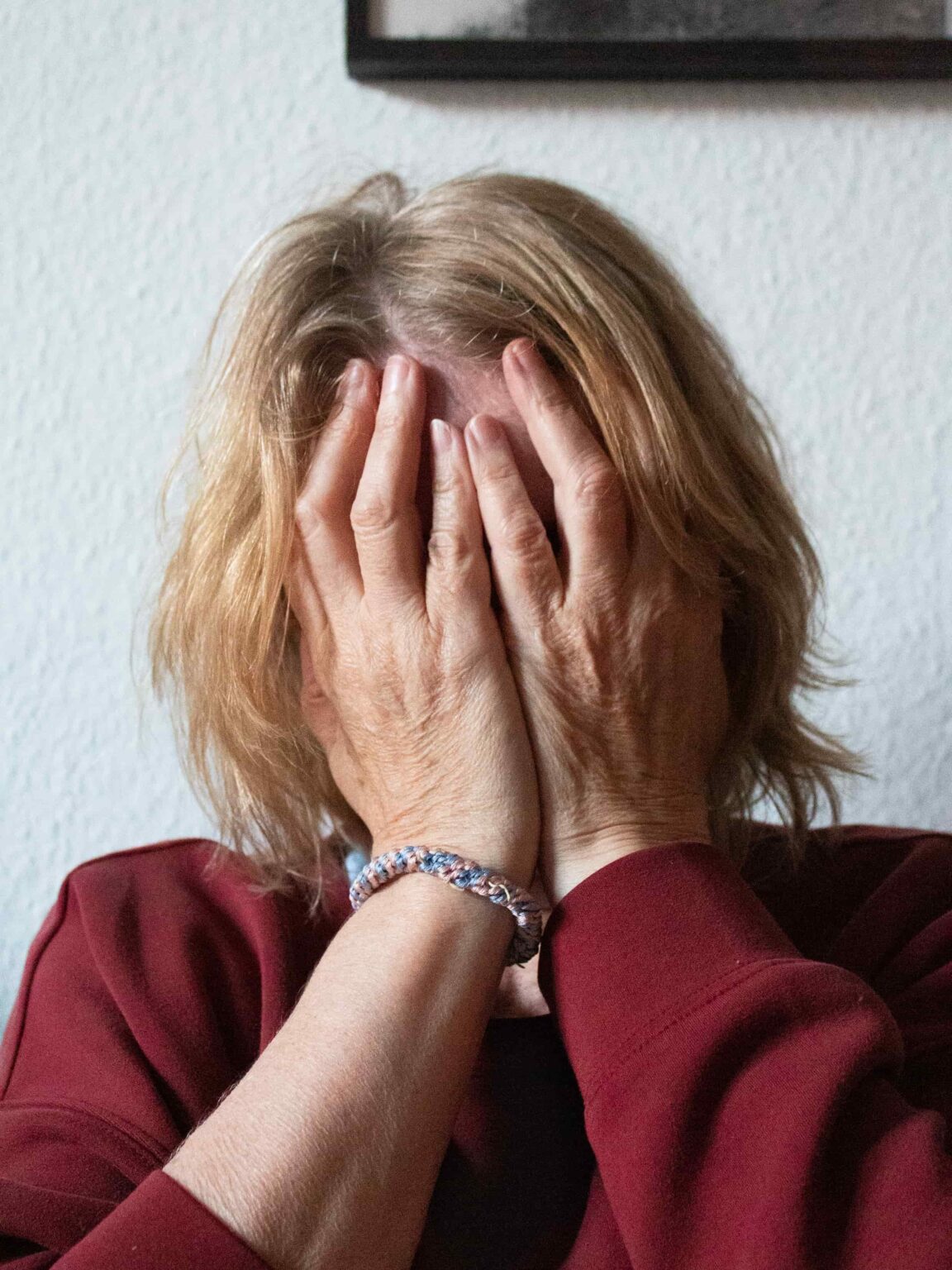 Eine blonde Person in einem bordeaux-farbigende Sweatshirt hält die Hände vor ihr Gesicht – ein Werk der Fotoserie der Fotografin Charlotte Wagner